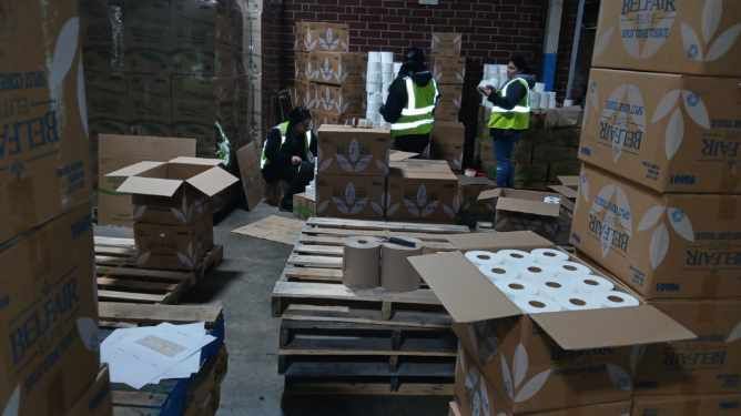 People in a warehouse sorting supplies, including boxes and toilet paper. Some wearing vests, boxes stacked on pallets.