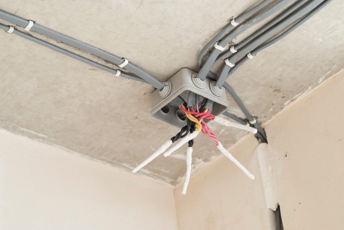 Electrical wiring junction box on a concrete ceiling with gray cables, white conduit, and exposed wires.