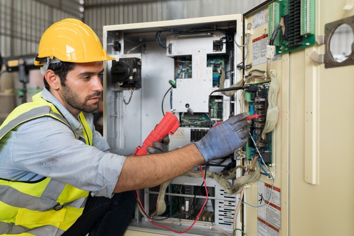 Male electrician in hard hat tests wires in an electrical panel. Yellow vest, blue gloves.