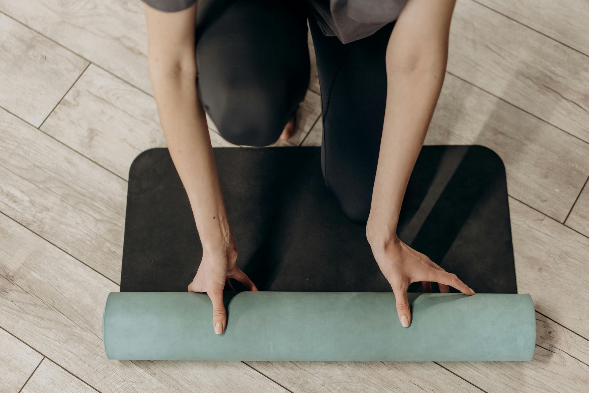 A white person in yoga leggings is rolling a green and black yoga mat over a wooden floor