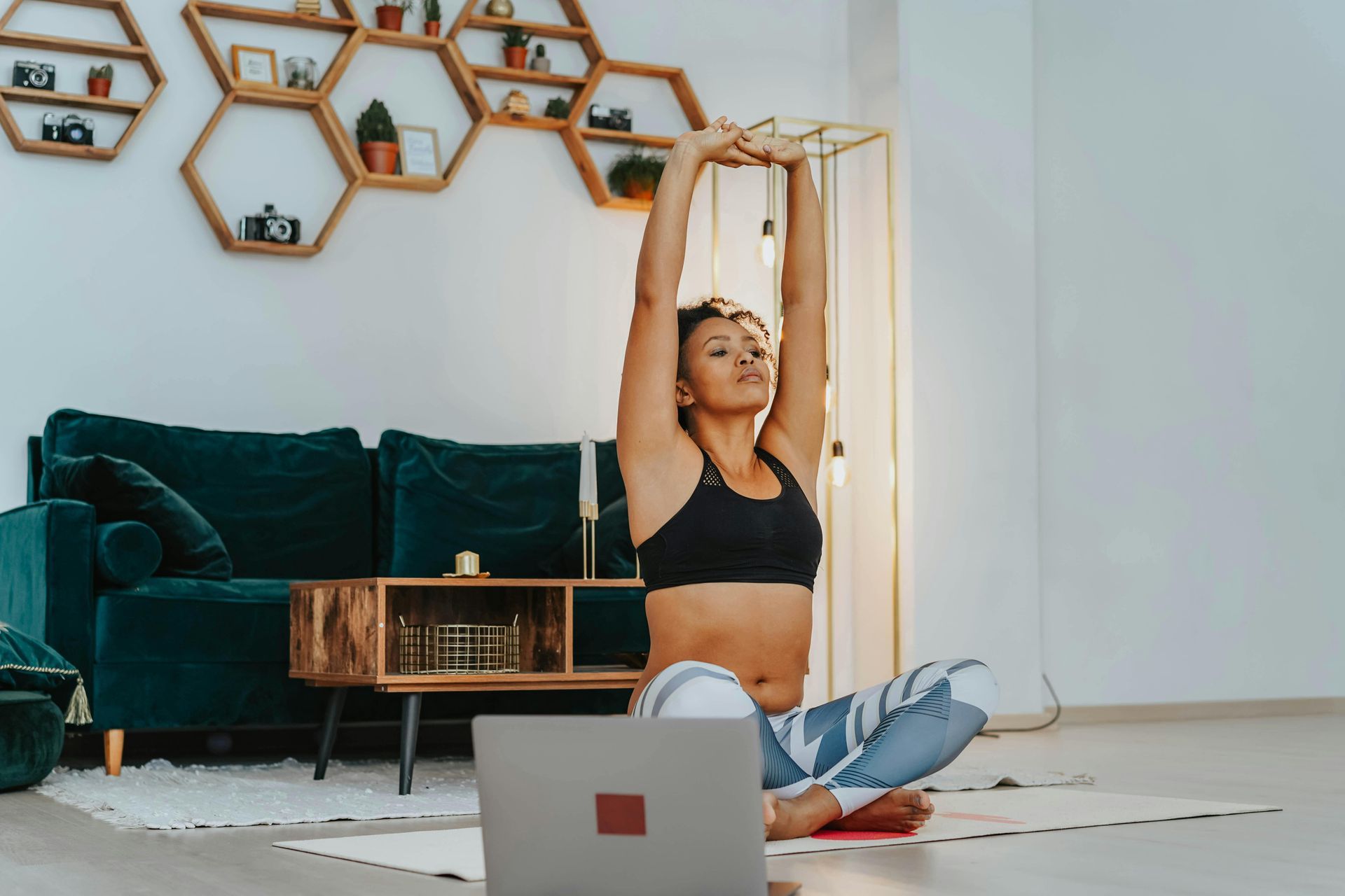 A Black woman in yoga leggings and a sports top stretches her arms above her head, sat on a yoga mat in front of her laptop. She is sat in front of a sofa and coffee table.