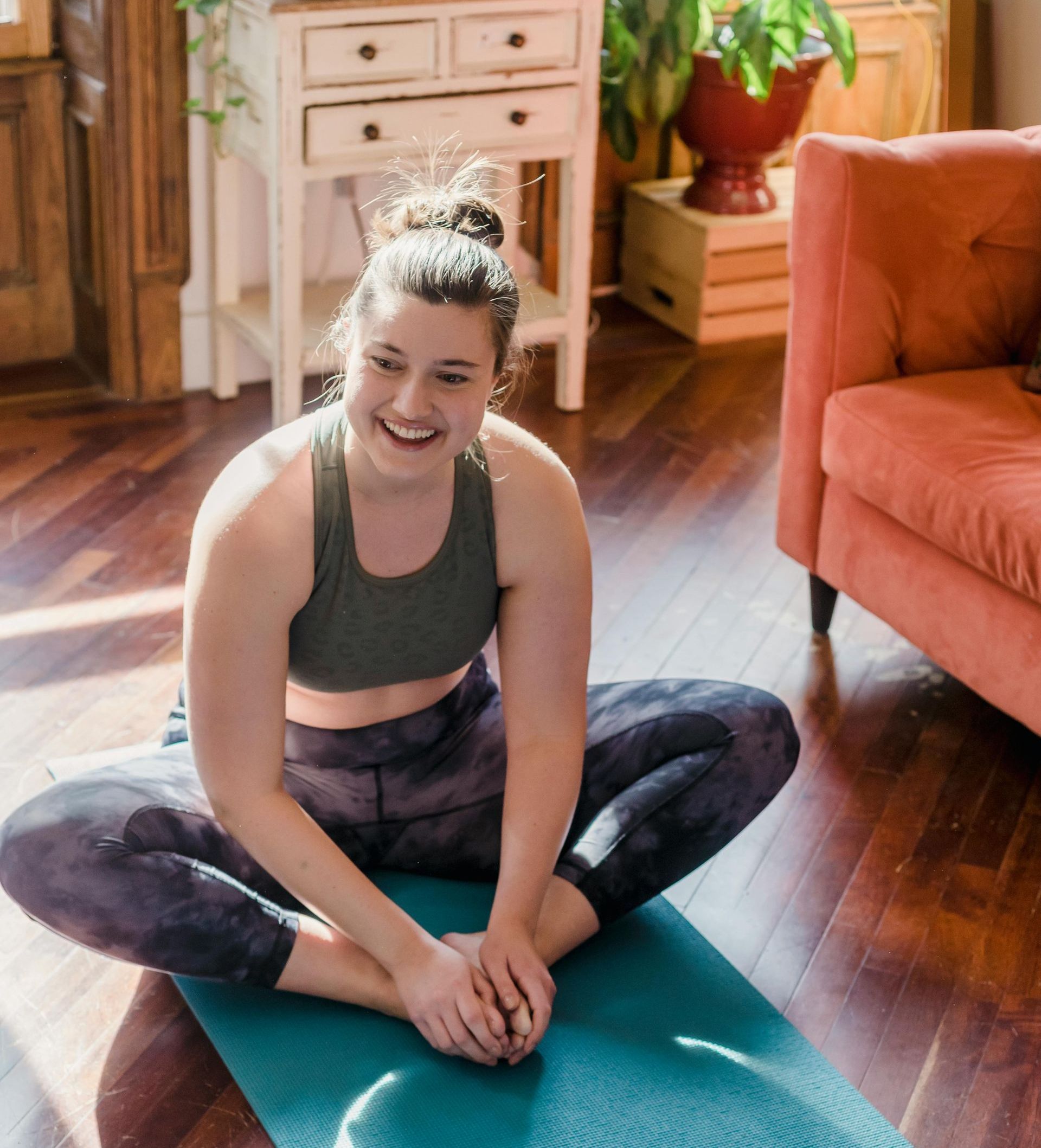 A white woman with he hair in a bun, wearing a sports top and yoga leggings, sits on a yoga mat in front of a sofa. She is smiling and holding her feet in a seated position.