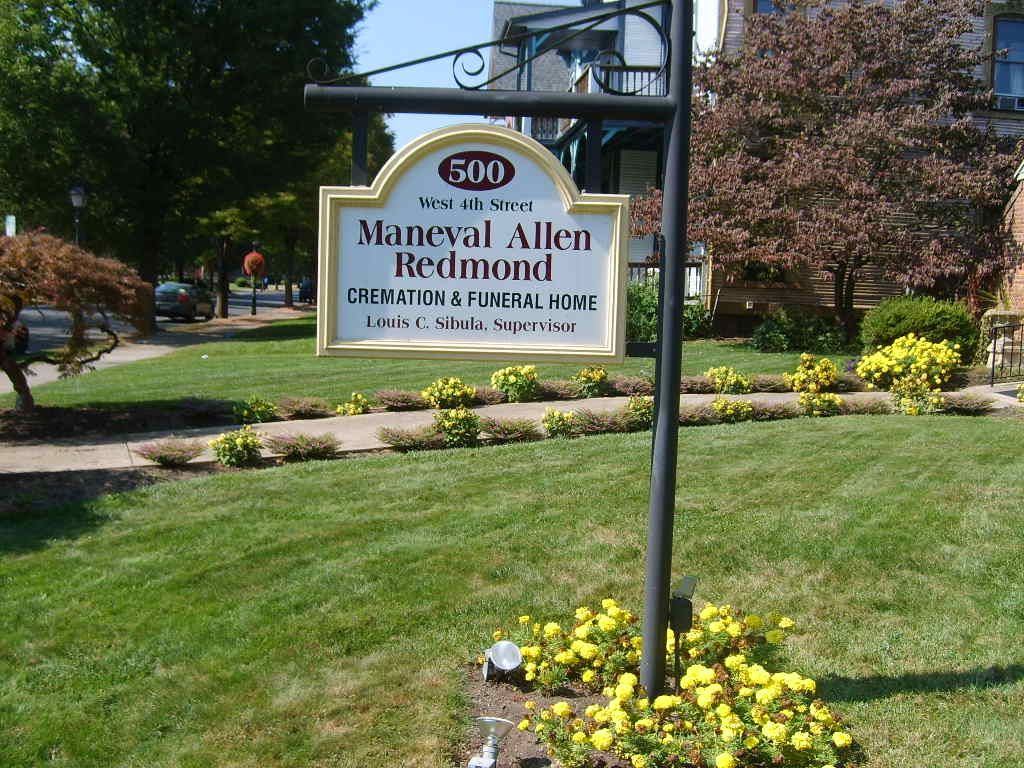 Sign for Maneval Allen Redmond, a funeral home, on a green lawn with yellow flowers.