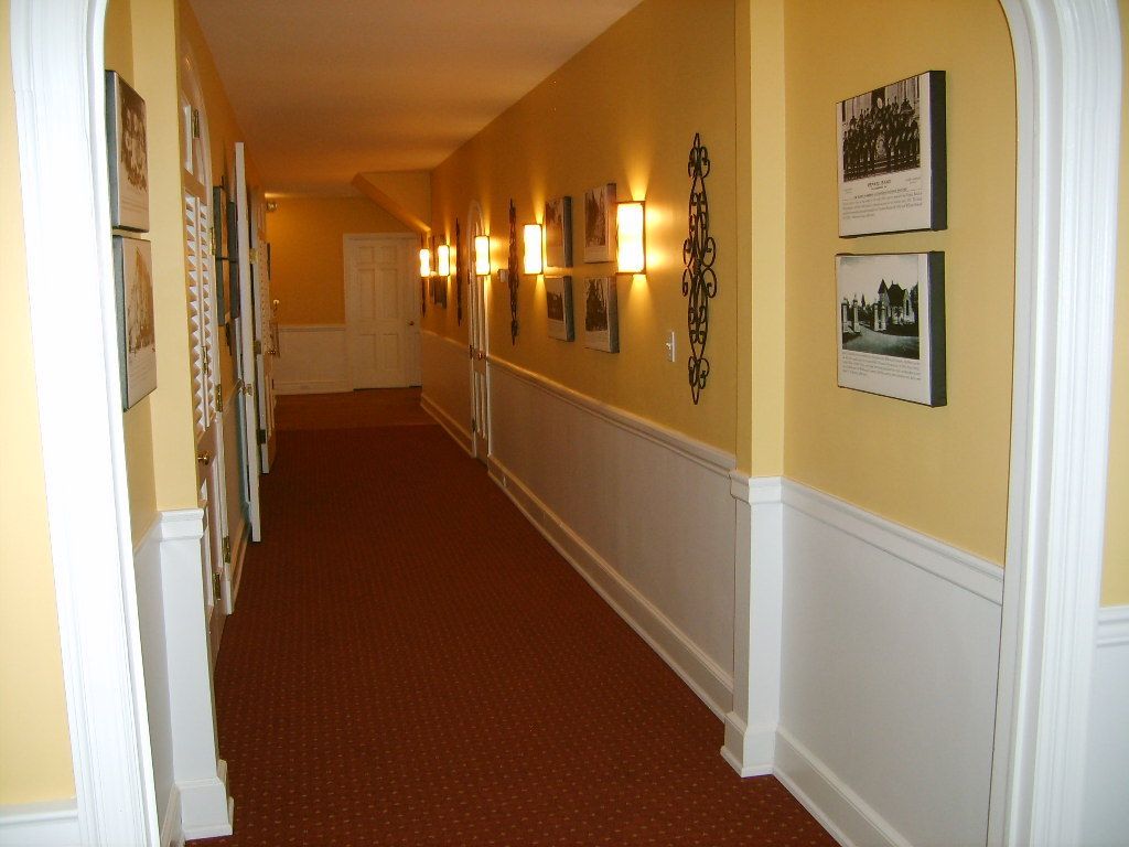 Long hallway with yellow walls, brown carpet, white wainscoting, and photos.