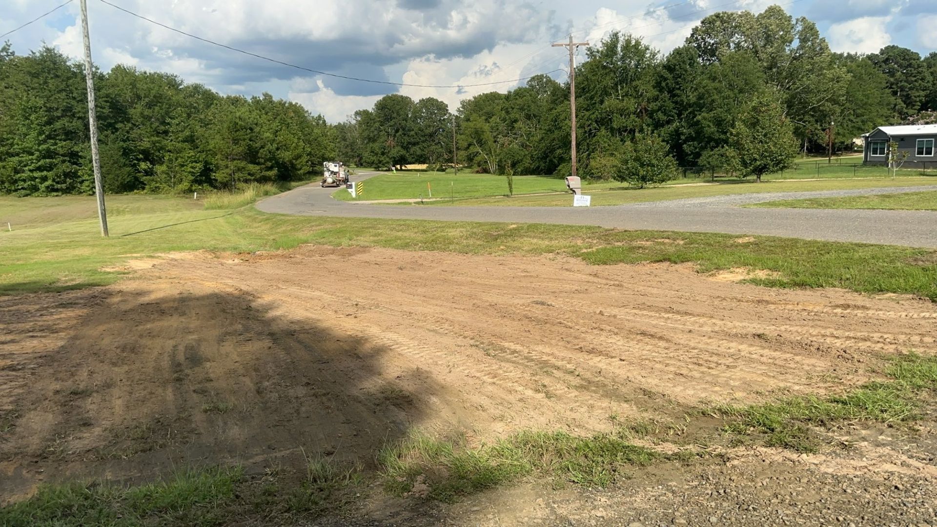 Dirt patch and paved road lead to trees under cloudy sky.