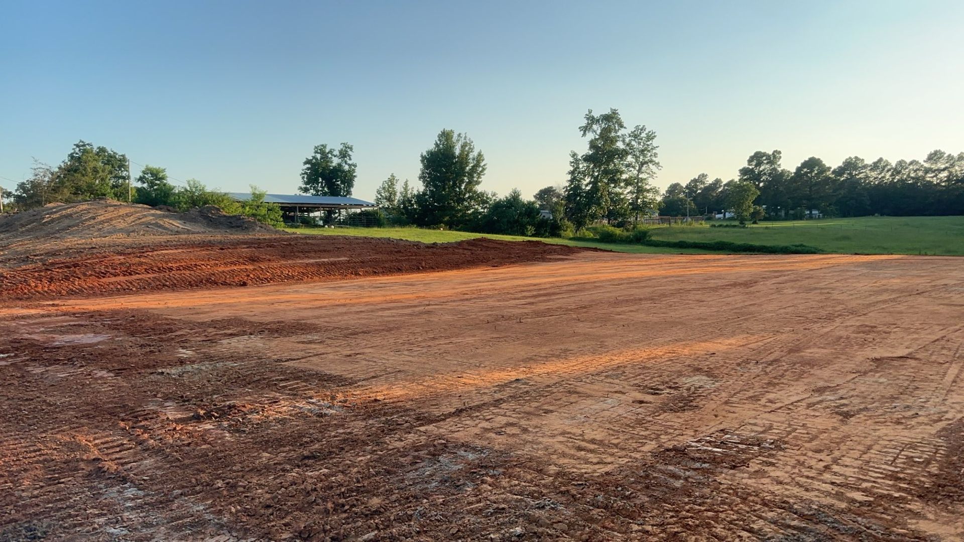 A barren construction site with reddish soil, green trees, and a building in the background under a blue sky.