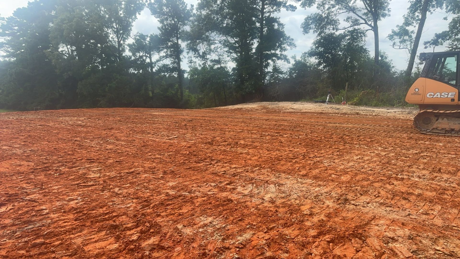 Red earth clearing with an excavator, trees in the background.