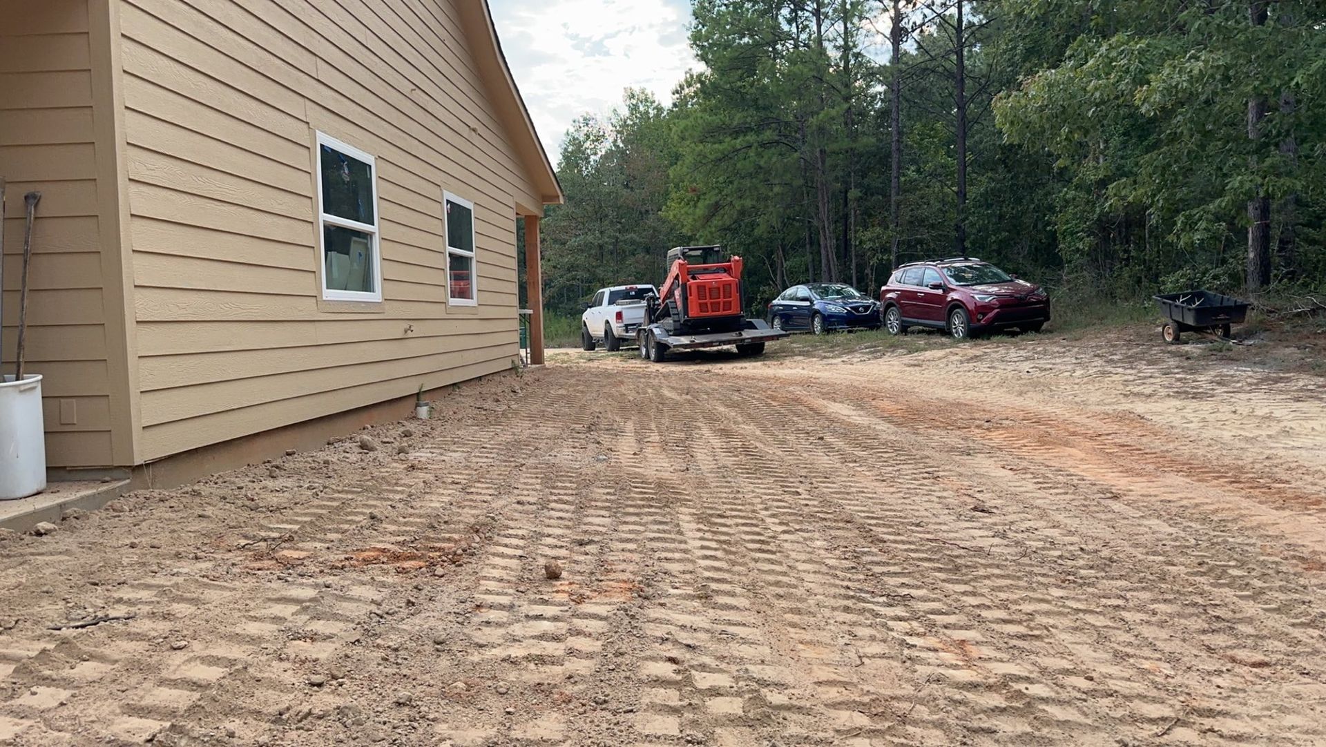 A house under construction with construction equipment on a dirt driveway. Cars and trees are in the background.
