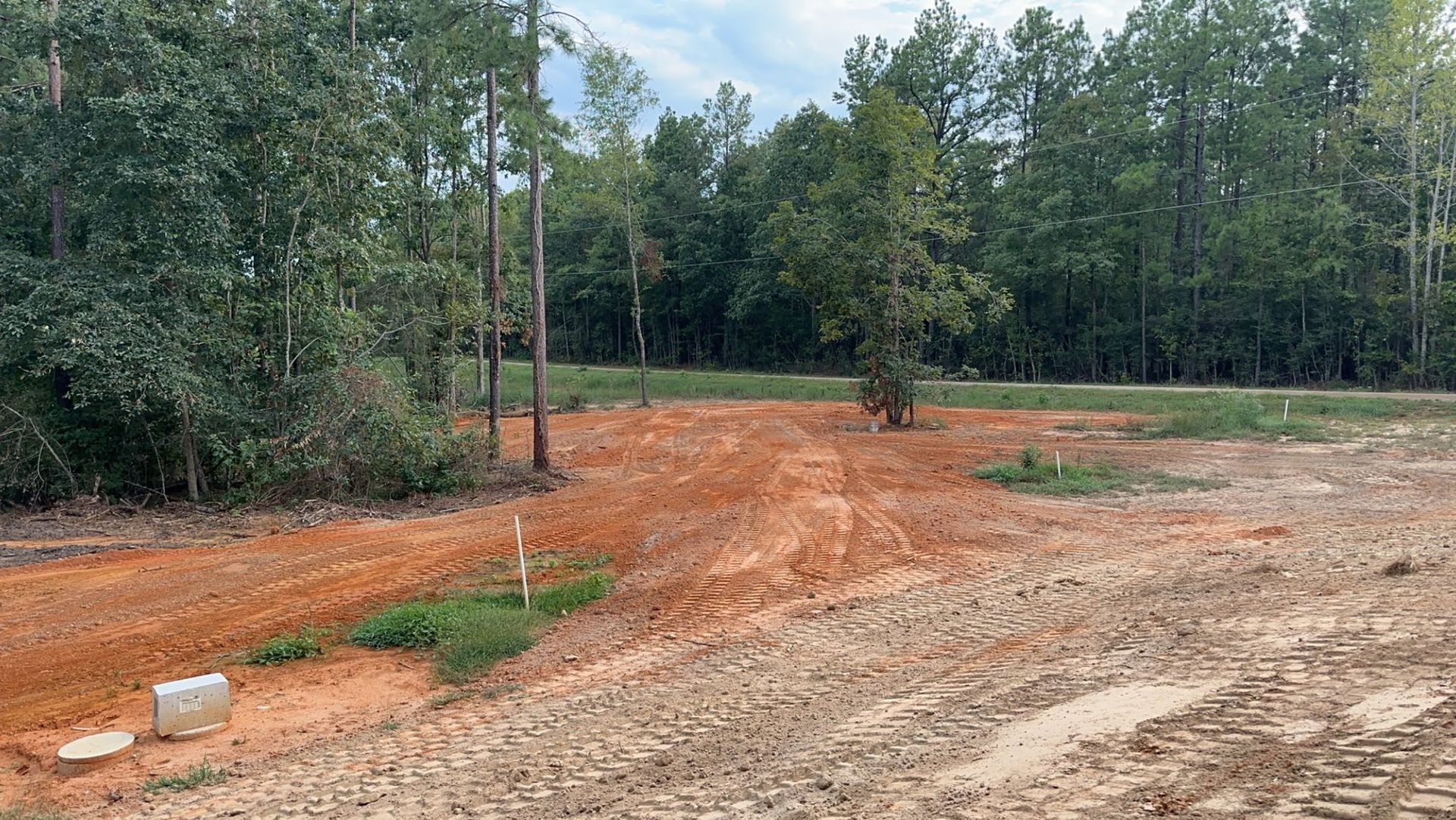 Dirt lot with power lines and trees in a wooded area, with some grass and a utility box.