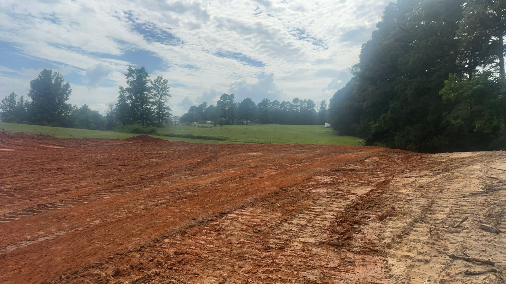 Red dirt foreground, grassy field in the distance, trees on the right, cloudy sky.