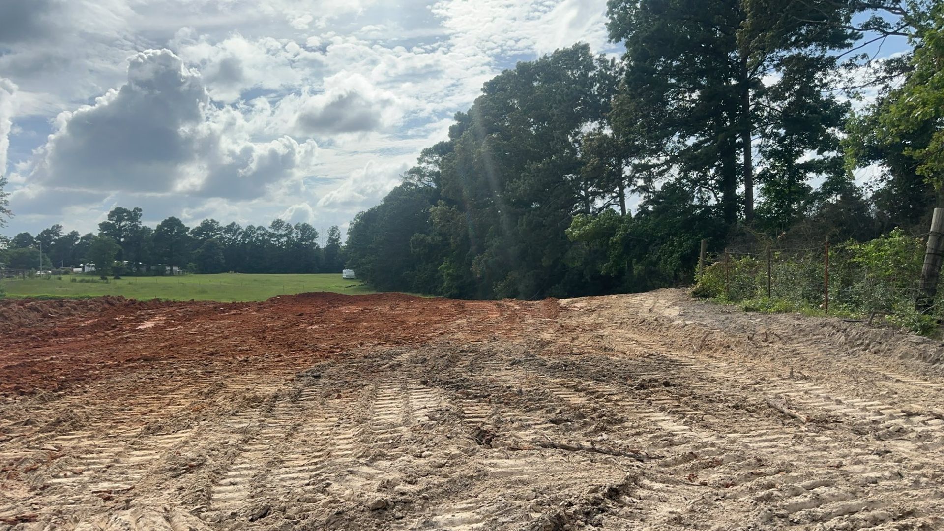 Muddy construction site with red clay, adjacent to a field and a line of trees under a cloudy sky.