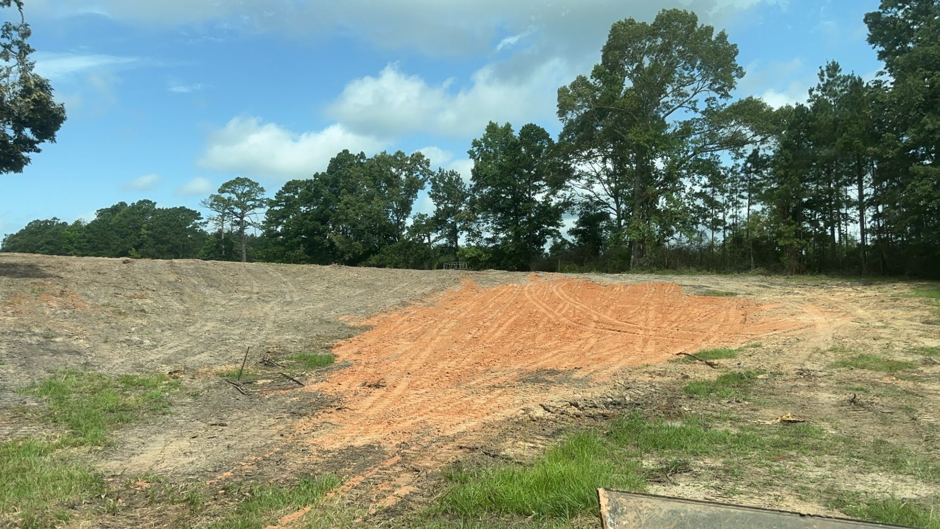Dirt pile on bare land with green grass, trees, and a partly cloudy sky.
