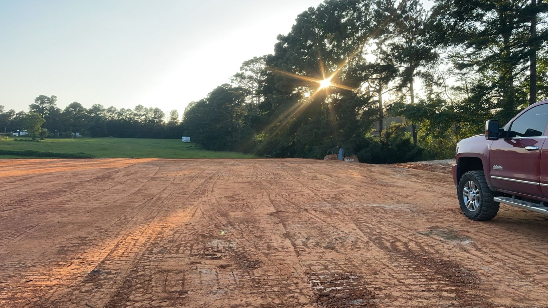 Red dirt construction site with a pickup truck, sunlight through trees.