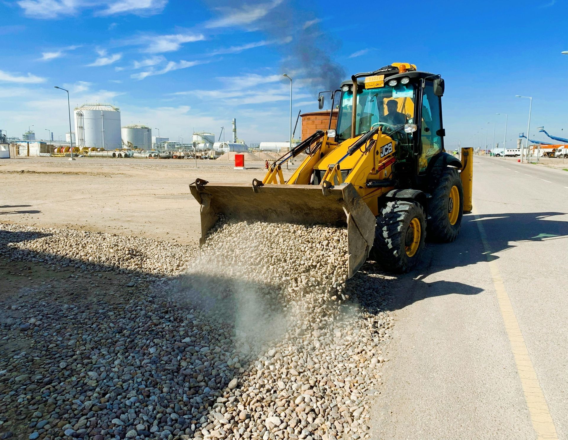 Yellow backhoe spreading gravel on a paved road at a construction site.