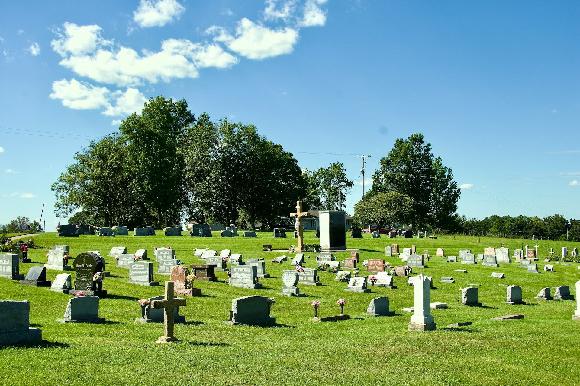 Sunny cemetery with rows of headstones, green grass, and trees under a bright blue sky 