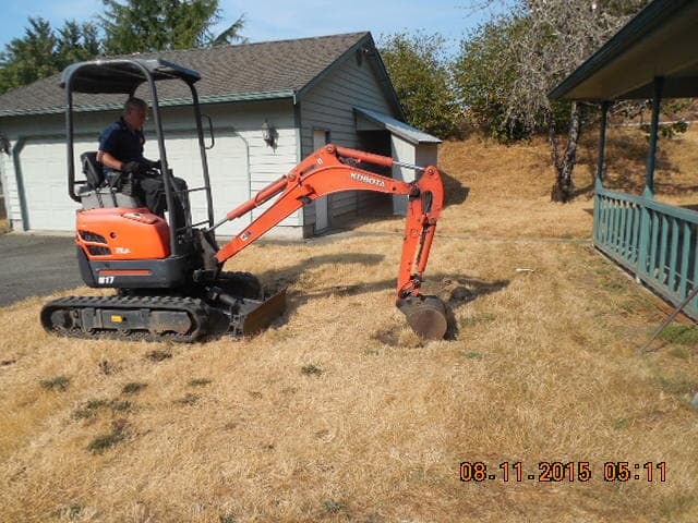 Man operating a small orange excavator on dry grass near a garage and house on a sunny day.