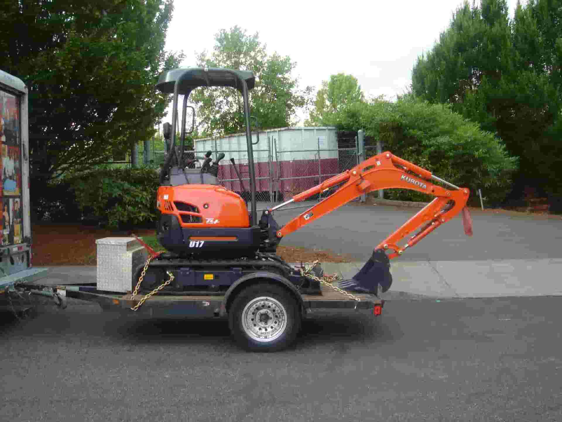 Orange mini-excavator secured on a trailer, parked on asphalt, near a tree line and a truck.
