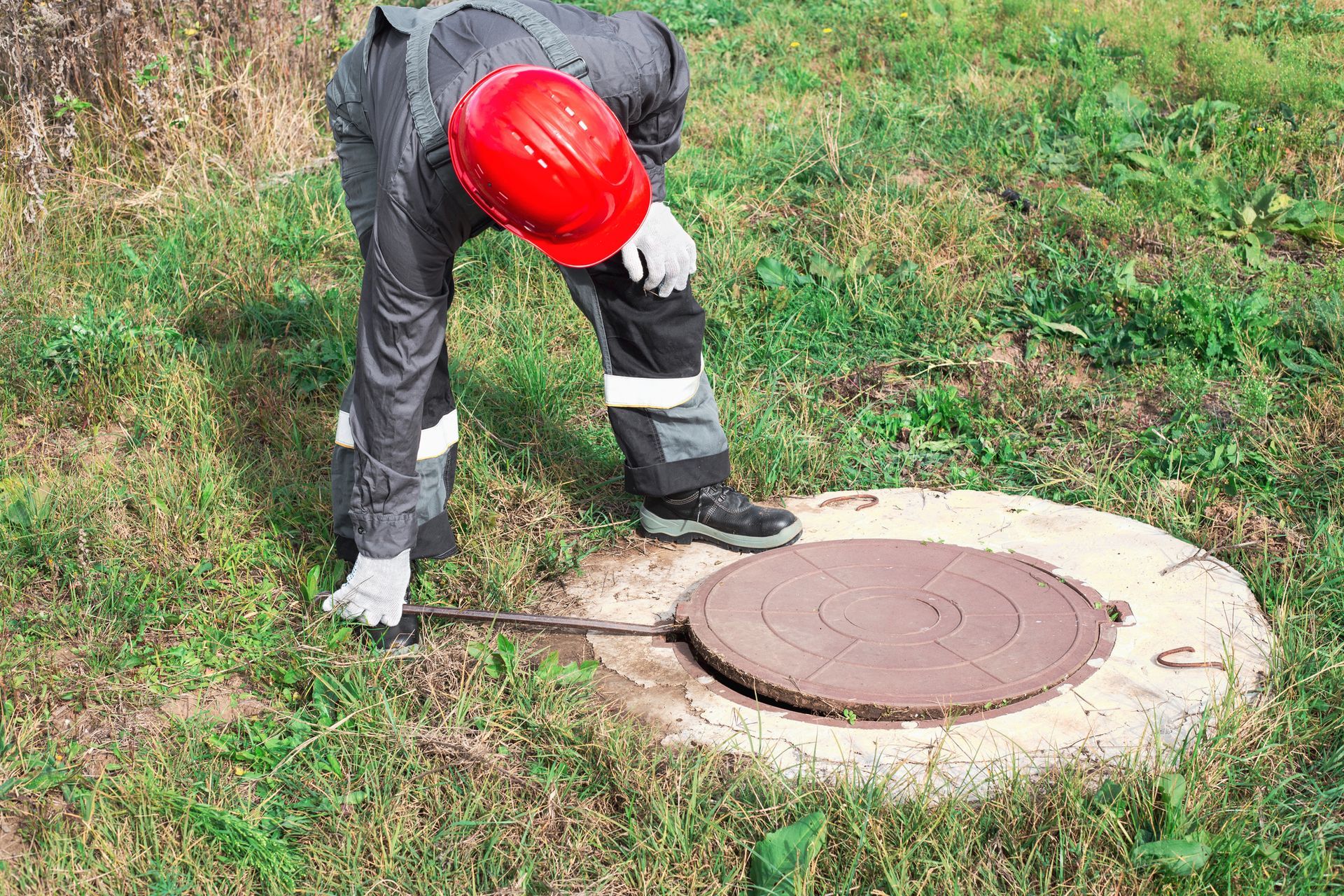 A plumber, wearing a red helmet, opens a manhole with a crowbar to perform septic system repair. 
