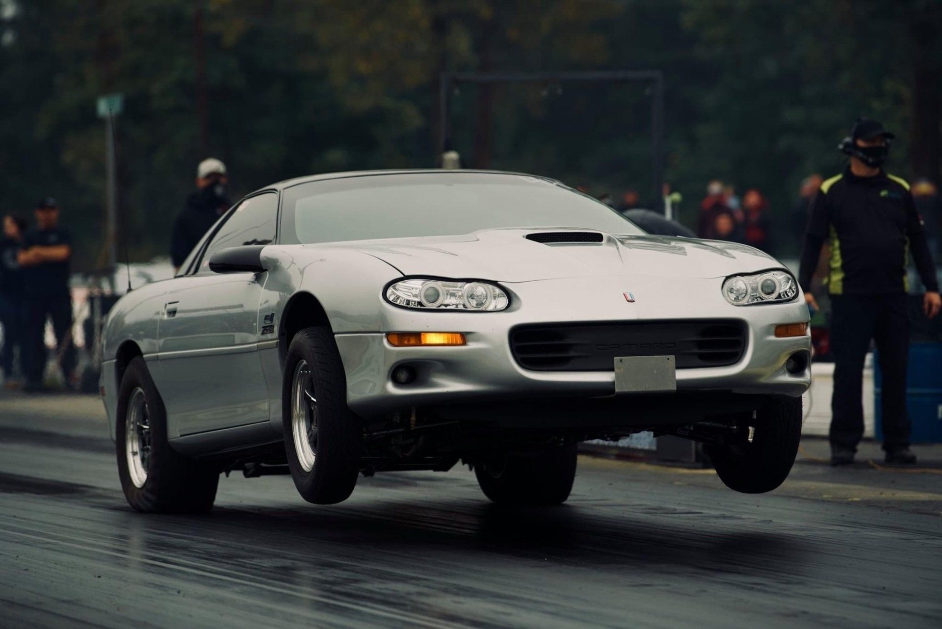 Silver Camaro drag racing, front wheels off the ground on a track.