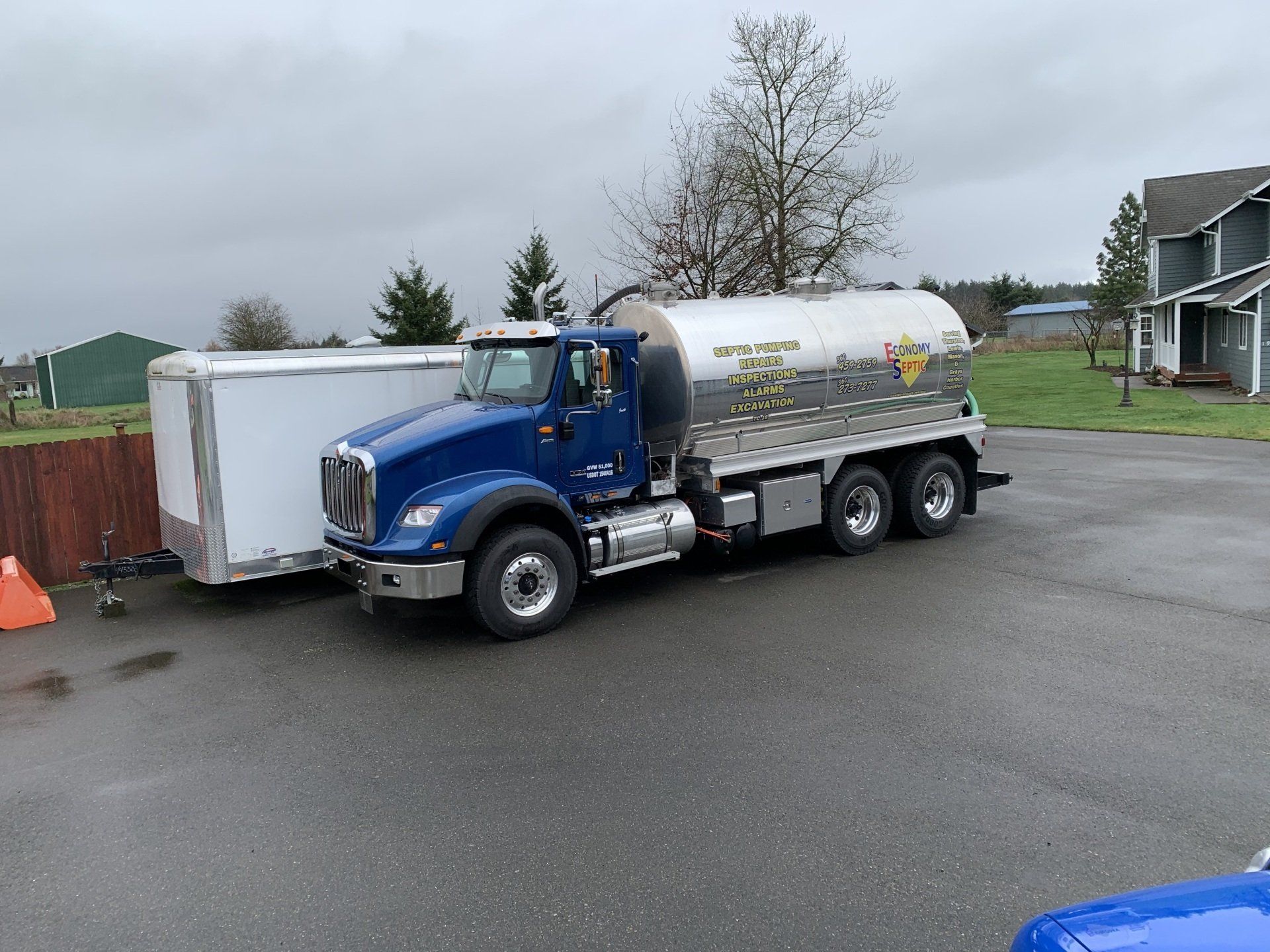 Blue tanker truck parked near a white trailer on a paved driveway in front of a house on a cloudy day.