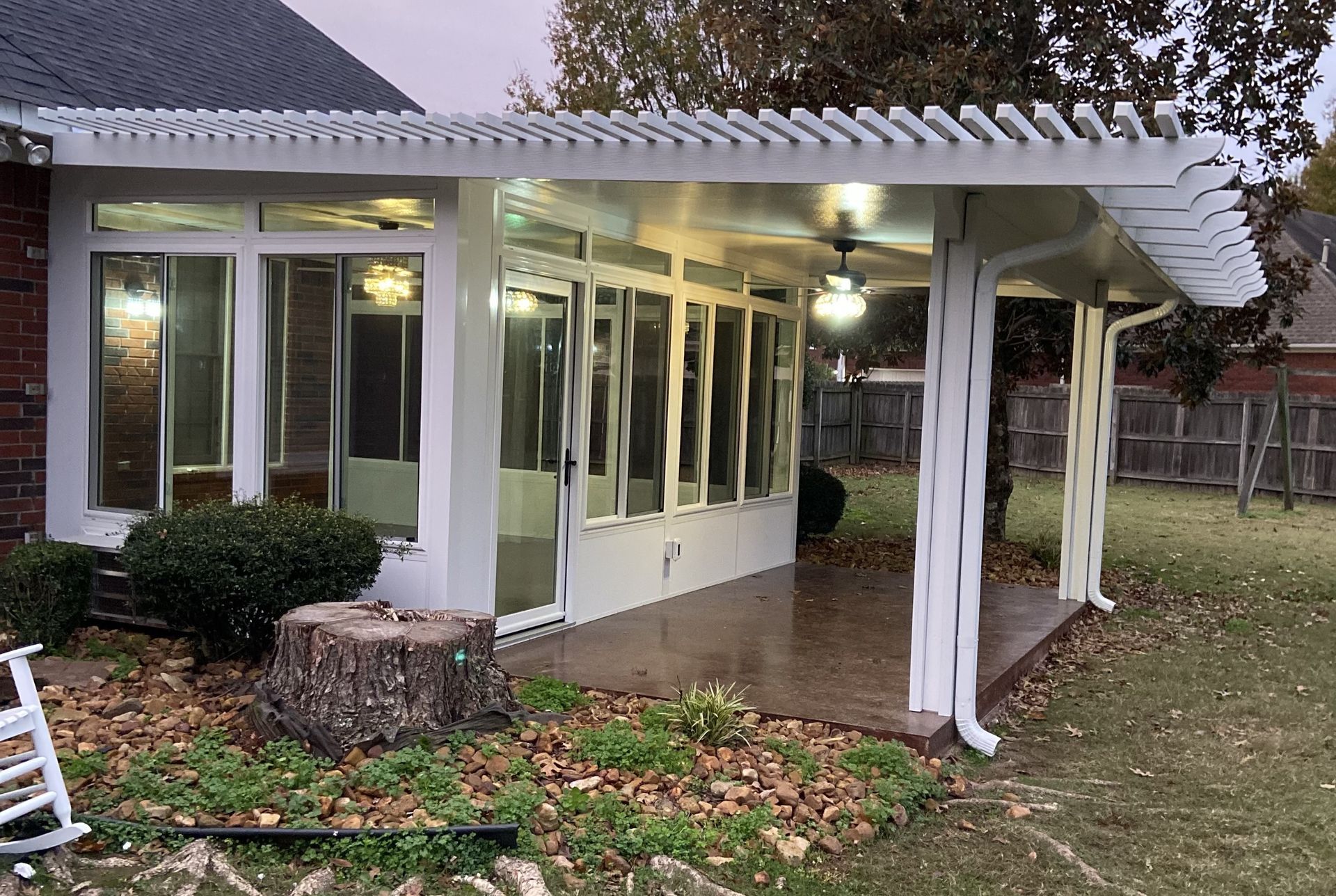 Sunroom with white frame, glass windows, and pergola-style roof attached to a house. Exterior with tree stump and yard.