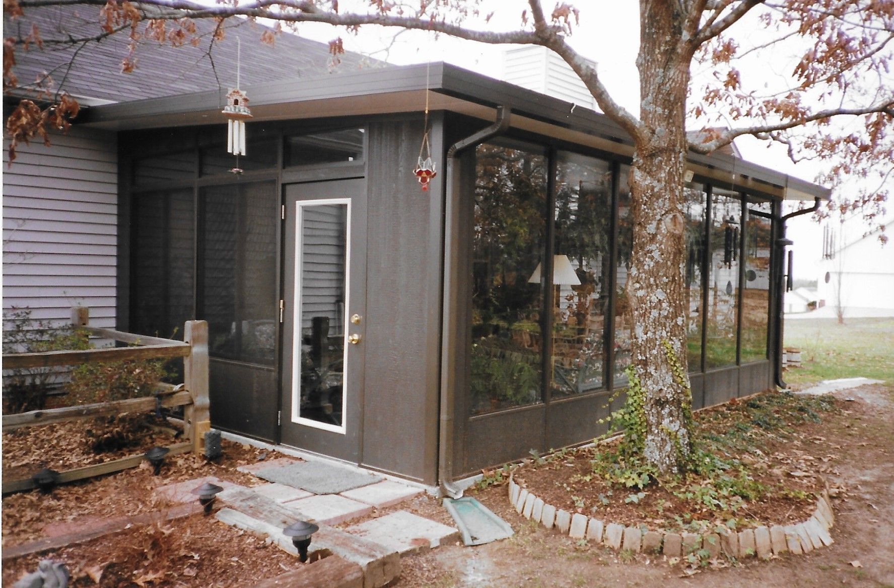 Brown sunroom addition with large windows, next to a tree and a house with a gray exterior.