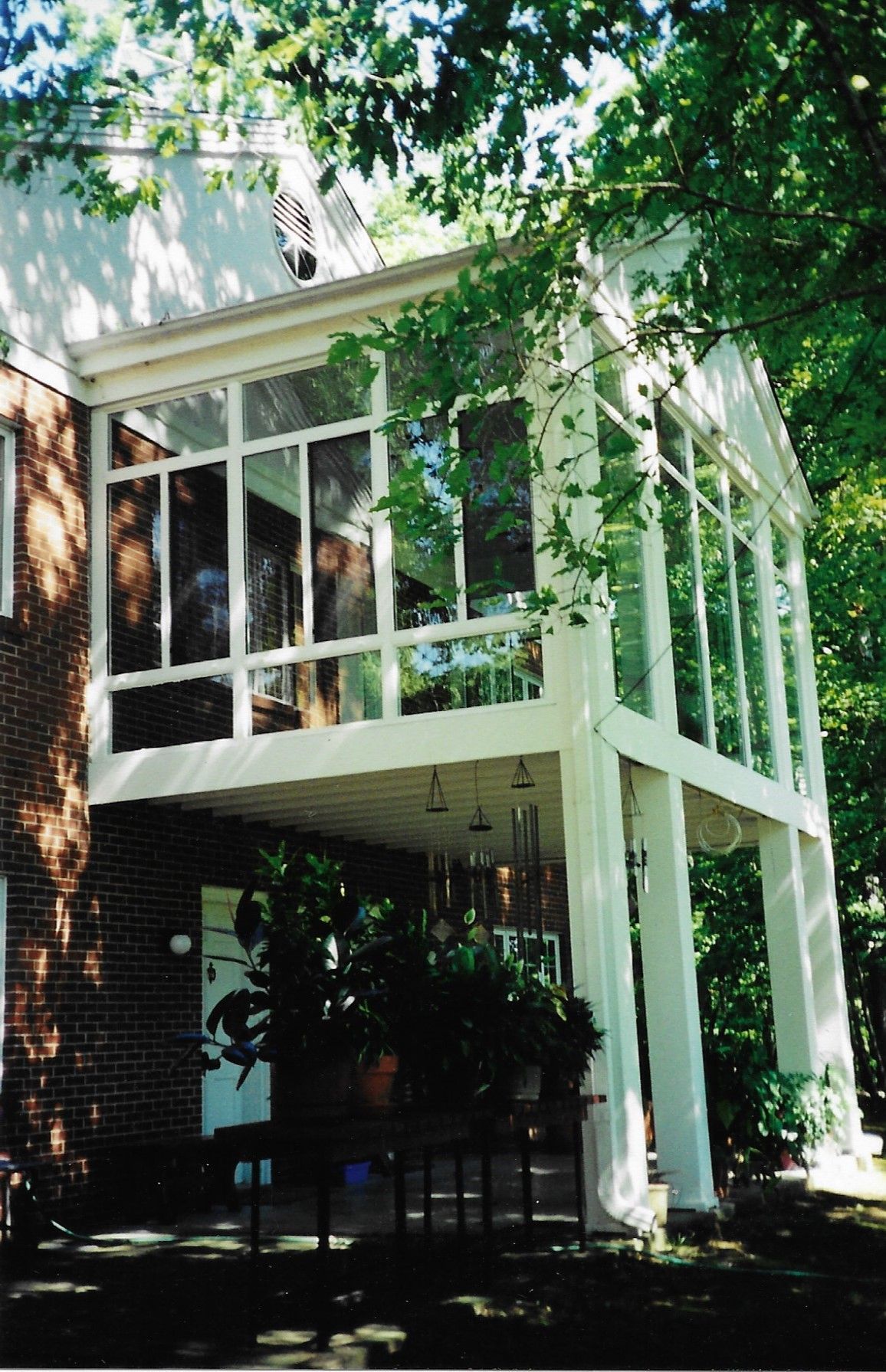 White-framed sunroom with glass walls, attached to a brick building. Trees surround the structure, shaded patio below.