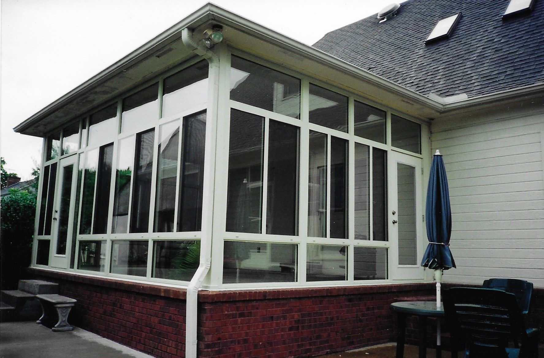 A sunroom attached to a house with brick base, glass windows, and a blue umbrella nearby.