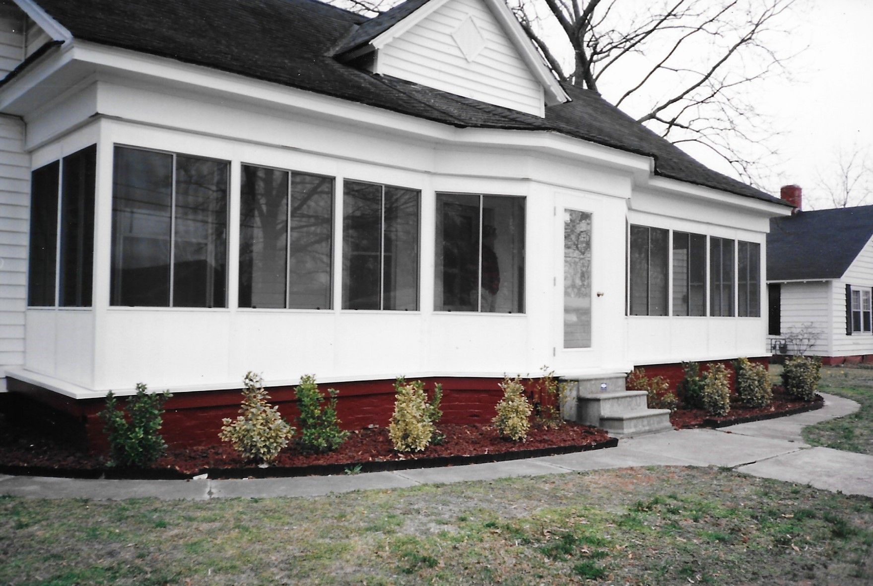 White house with screened-in porch, brick foundation, small bushes, and sidewalk.