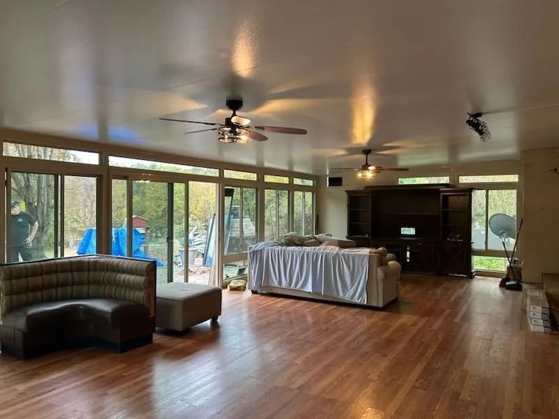 Spacious room with wooden floors, large windows, ceiling fans, furniture covered in white, and a sectional.