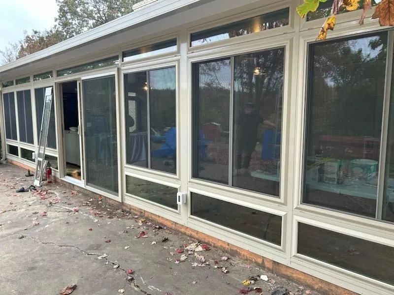 Glass-walled sunroom with open sliding door. Beige frames, dark lower panels, screen doors.