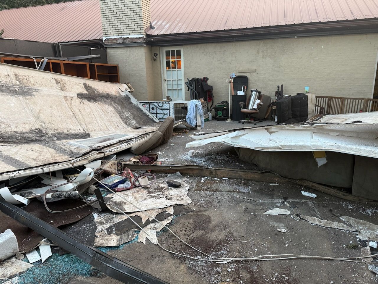 Debris-filled outdoor area; damaged roof, furniture, and objects scattered near a building with a white door.