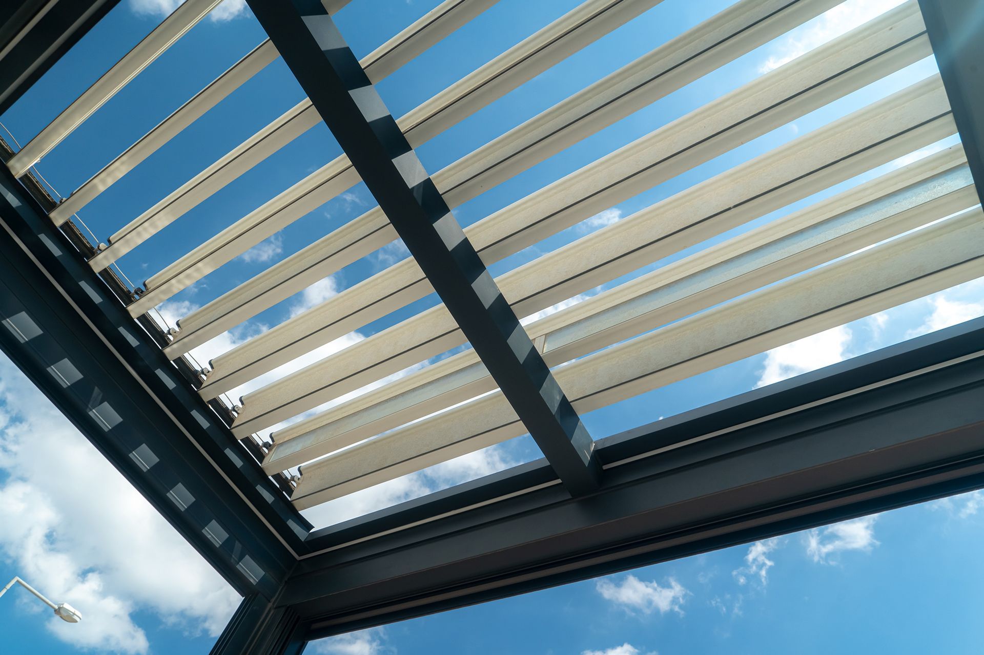 View upwards of a beige and aluminum pergola roof against a blue sky with clouds.