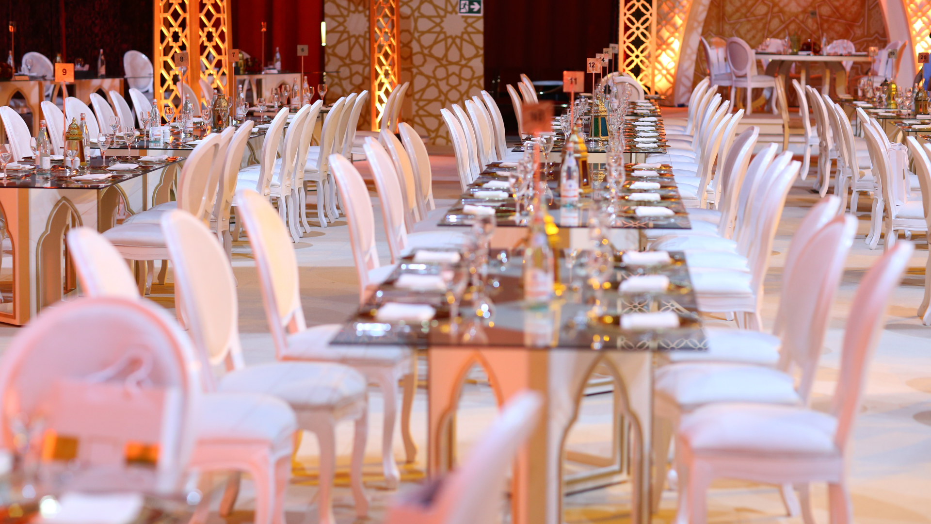 Elegant banquet hall with long tables, white chairs, and decorative lighting.