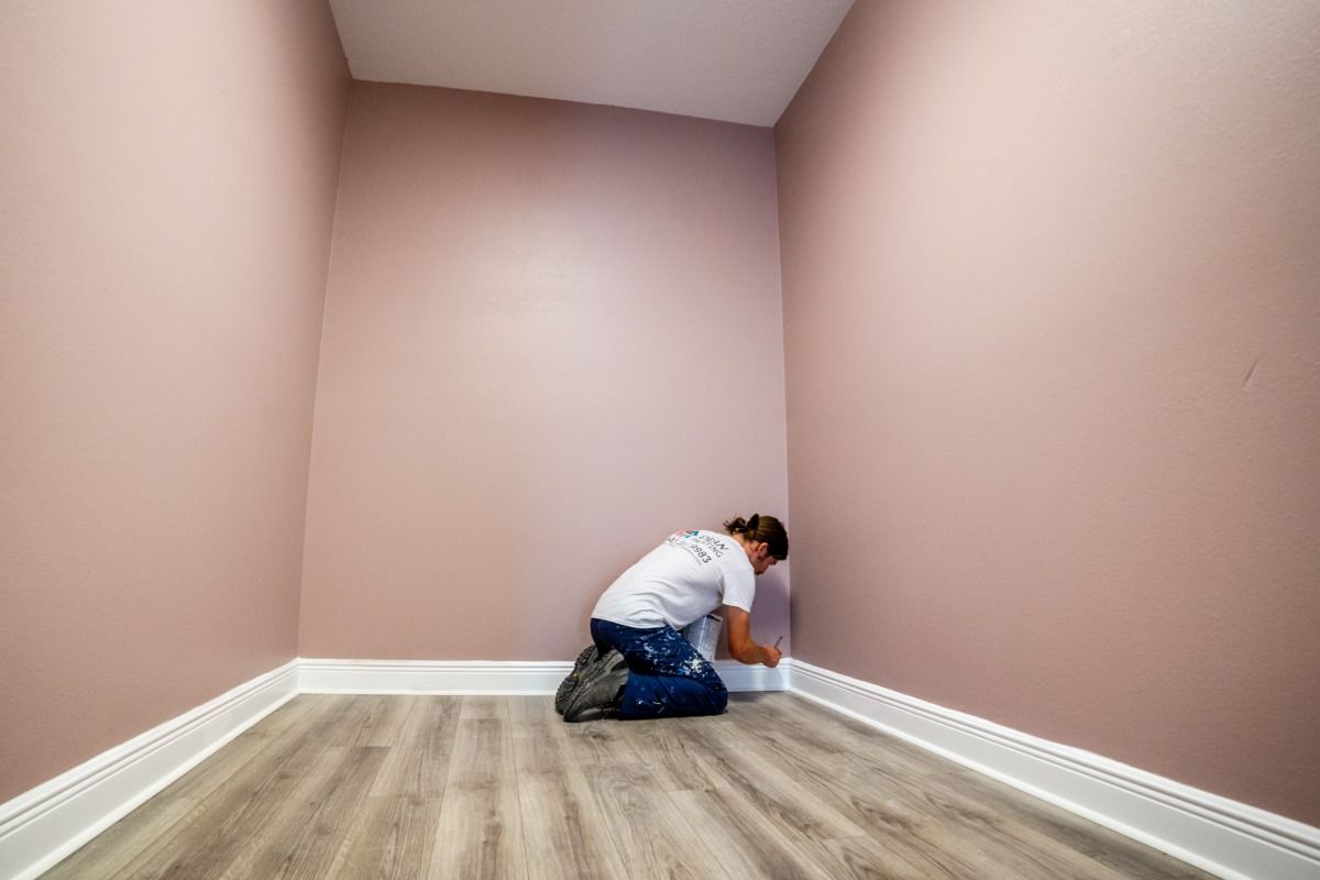 A man is kneeling down in a corner of a room painting a wall.