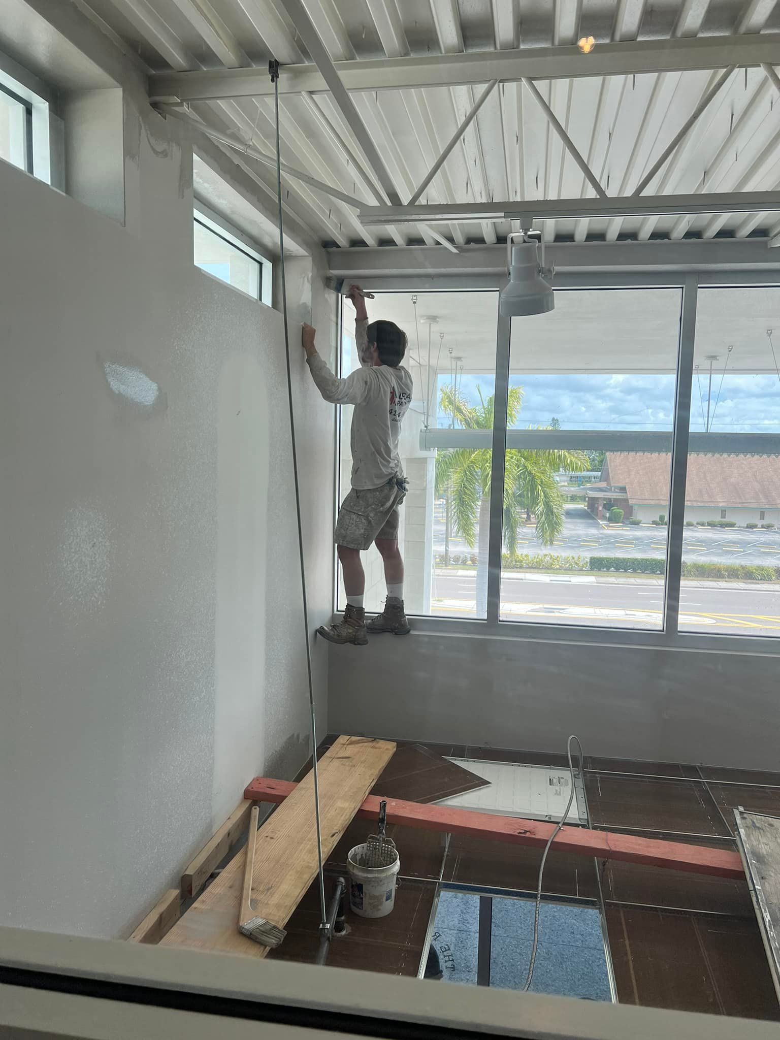 A man is painting a wall in a room with a lot of windows.