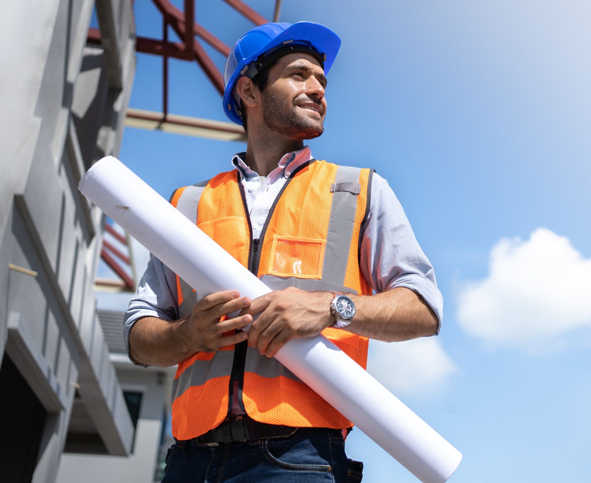 A smiling construction worker in a blue hard hat and orange vest holds blueprints at a building site against a blue sky.
