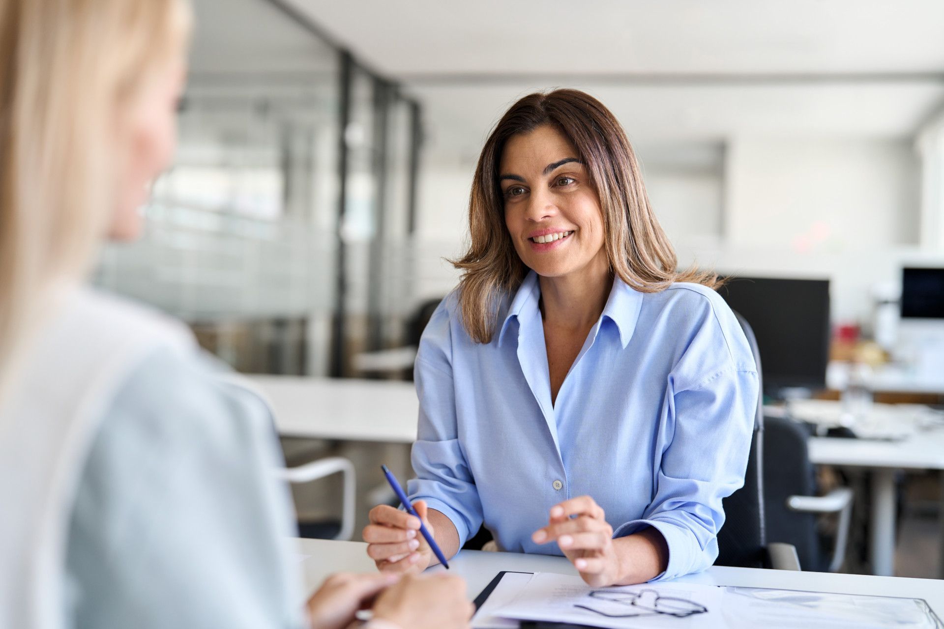 Two people sit at an office desk for a professional meeting, with one smiling while holding a pen during a conversation.