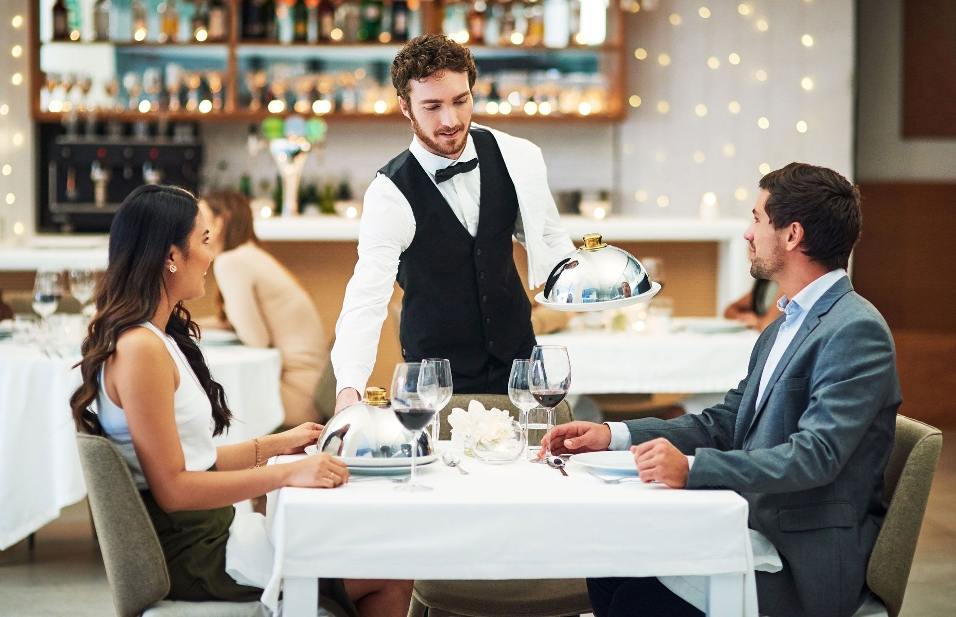 A waiter serves food to two people at a white-tablecloth table in a restaurant.