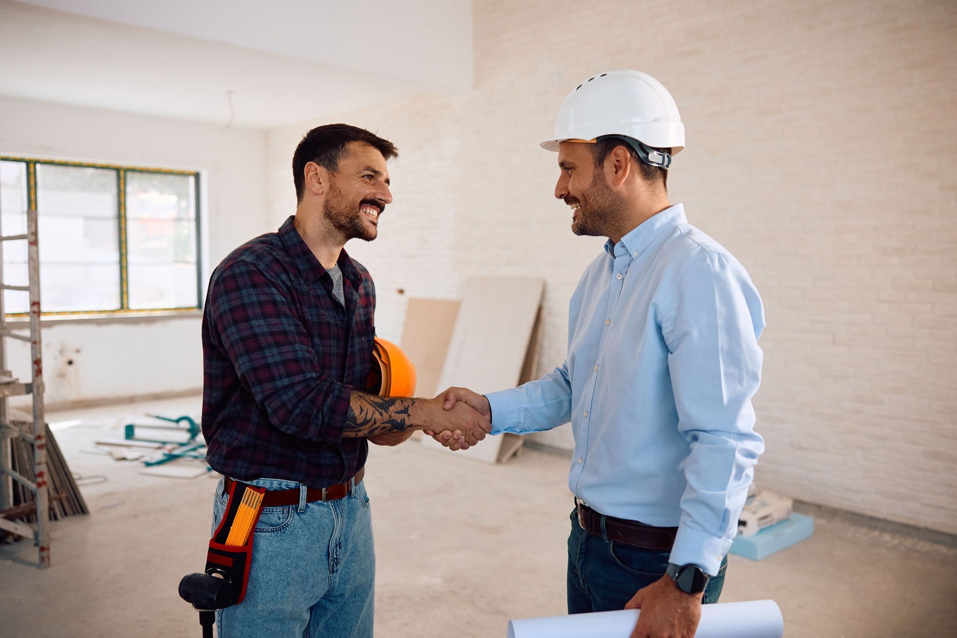 Two construction workers in a building under renovation shake hands and smile while facing each other.