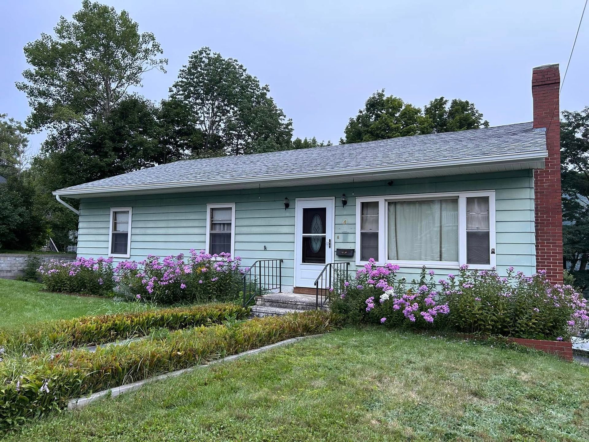 A blue house with black rain gutters, a brick chimney and flowers in front of it.