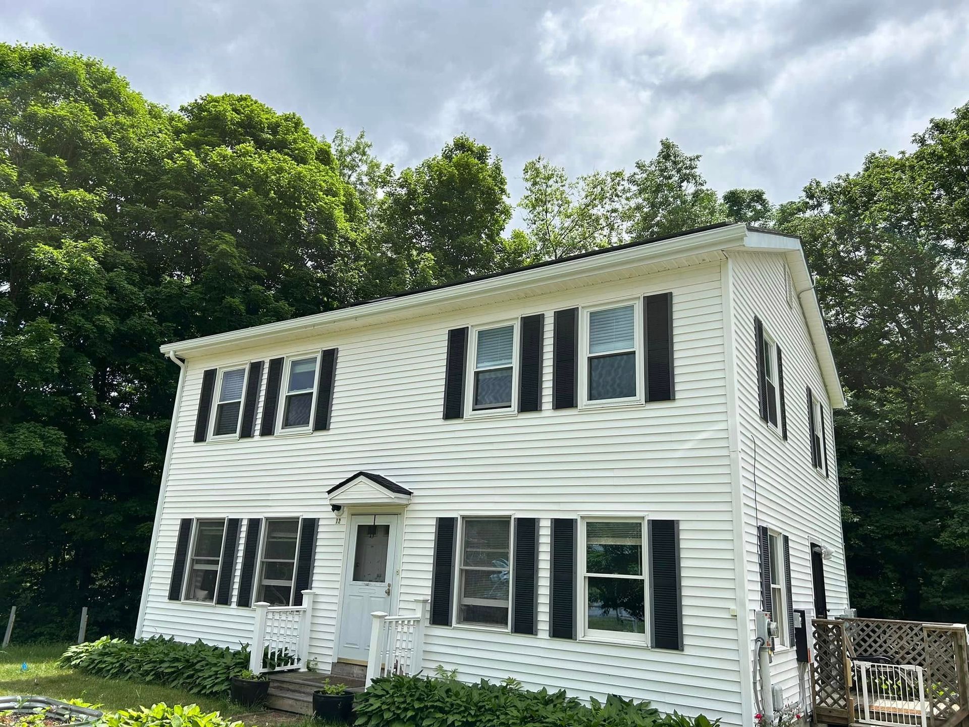 A white house with black shutters is surrounded by trees on a cloudy day.