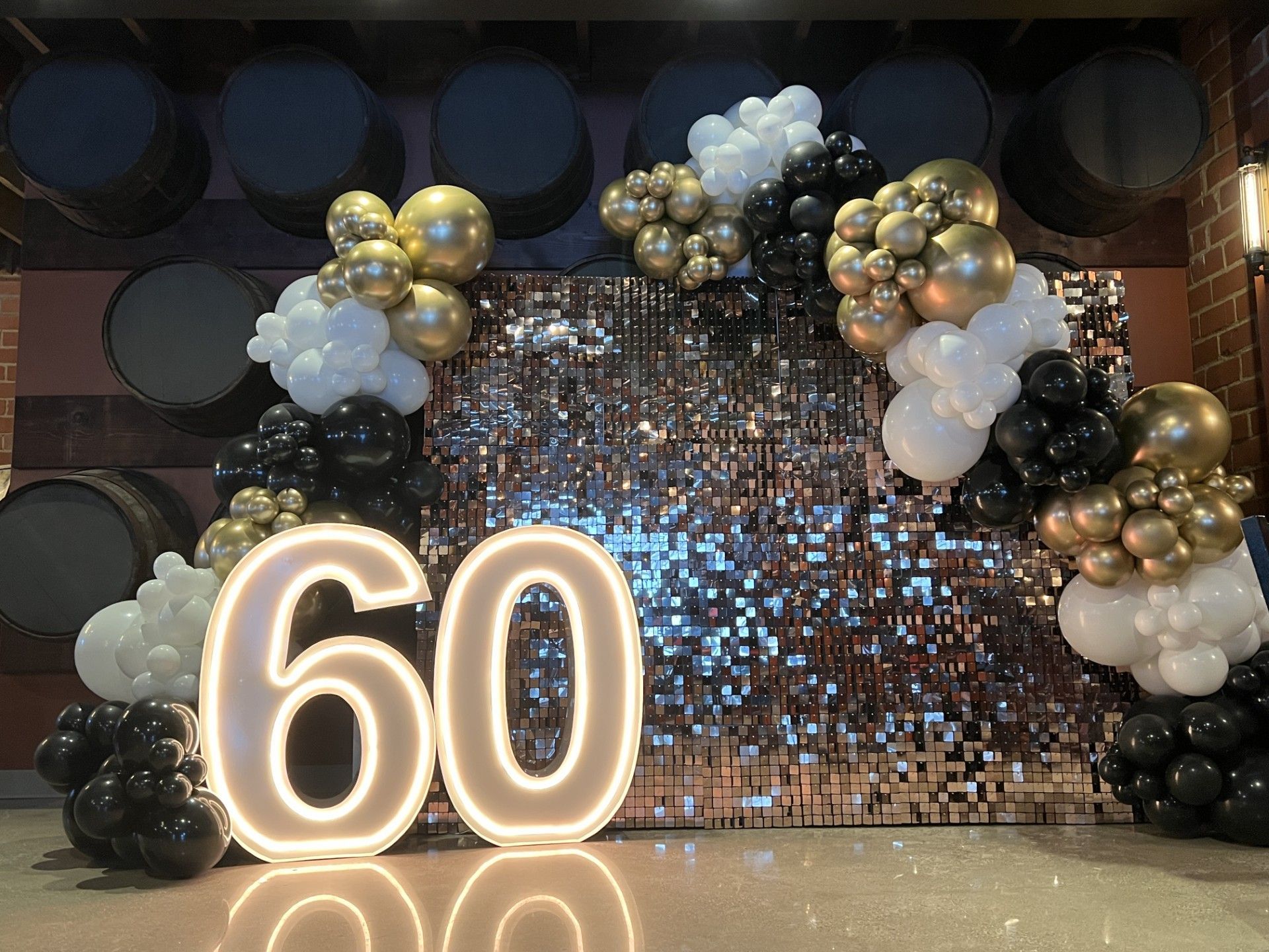 A luxurious 60th birthday celebration backdrop featuring a black, white, and gold balloon garland   elegantly framing a shimmering sequin wall.  The display is enhanced with large illuminated
