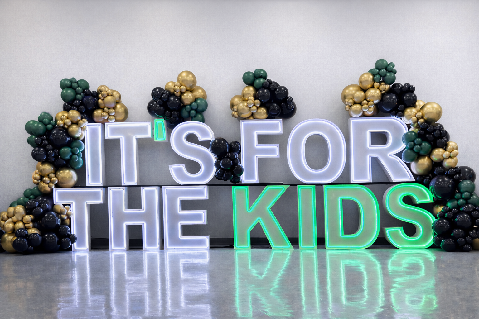 LED marquee letters spelling “It’s For The Kids” with emerald green, black, and gold balloon garland backdrop for a fundraiser event display by Designs by Jo Jo’s Creations serving Akron, Canton, and Cleveland.
