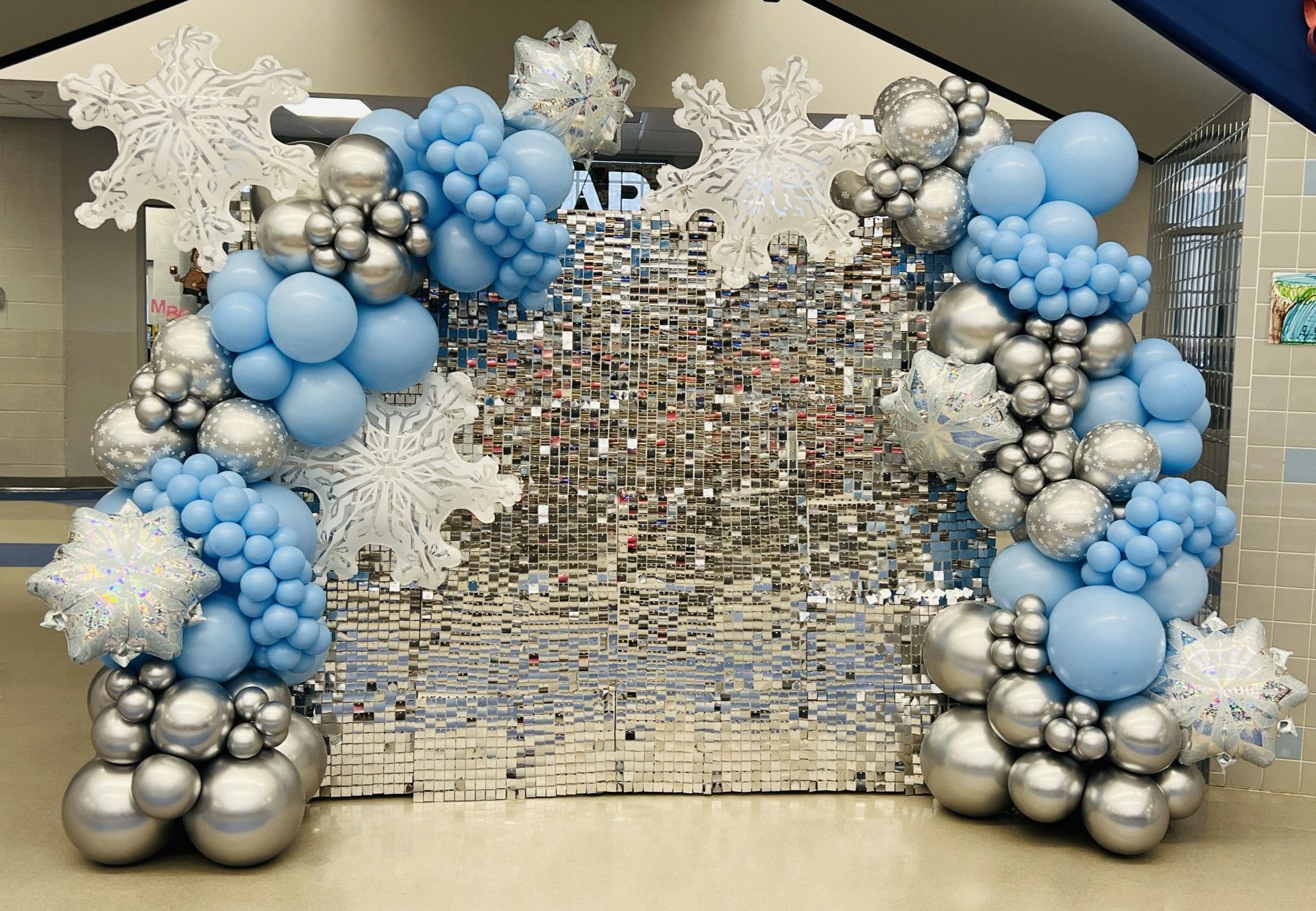 A winter wonderland-theme balloon backdrop featuring an elegant silver and light blue balloon garland framing a shimmering sequin wall.  The design is enhanced with large white snowflake cutouts and iridescent star-shaped balloons, creating a magical and frosty ambiance.  Perfect for winter events, holiday celebrations, Frozen-theme parties, or school dances, this setup adds a touch of glamour and seasonal charm to any occasion.