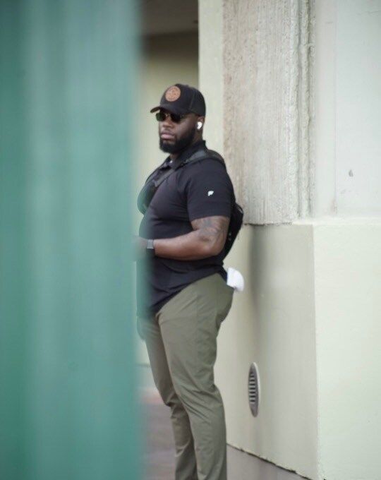A Black man in black shirt and cap, khaki pants, and earbuds stands outdoors near a wall.