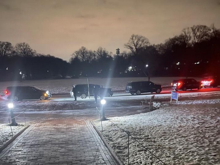 Vehicles parked on a snowy, dimly lit road at night. Trees in the background, bright pathway.