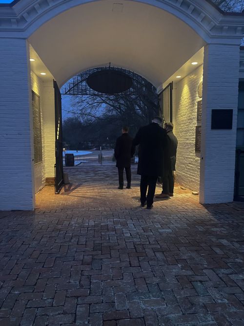 People stand under an arched gateway with lights. Night setting; brick pathway.