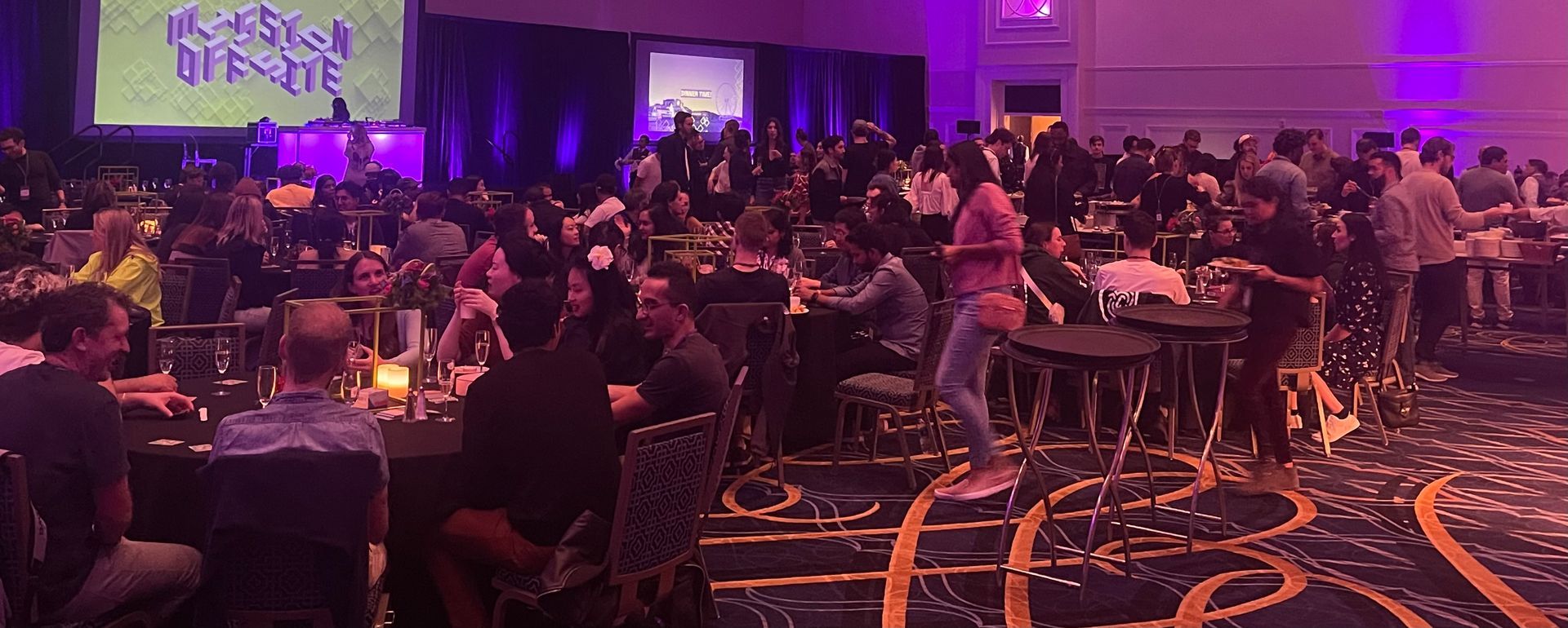 Large event with tables, people, and a stage with a screen illuminated in purple.