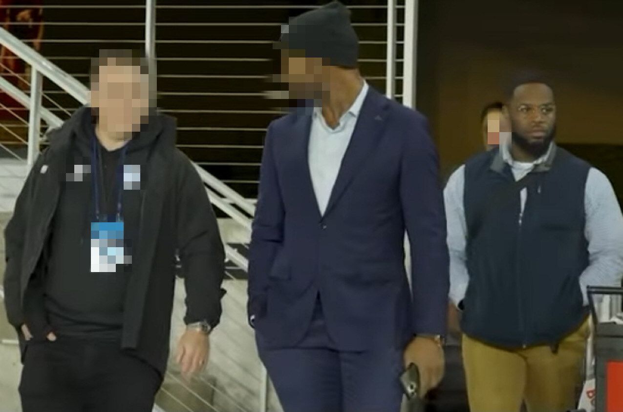 Three men walking: One in a suit, one in a vest, and one in a jacket, indoors at a stadium.